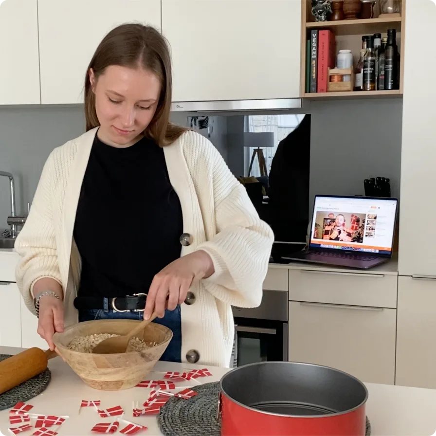 A learner baking in her kitchen with Danish flags on the counter while a Swap Language lesson plays on her laptop
