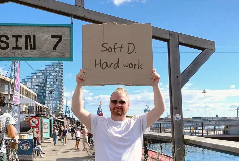 A Swap Language learner at a Danish waterfront holding a handwritten cardboard sign reading “Soft D. Hard work.”
