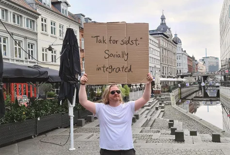 A Swap Language learner on a Danish canal street holding a handwritten cardboard sign reading “Tak for sidst. Socially integrated.”